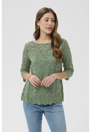 Kvinde med langt bølget hår iført en grøn blomsterblonde-trekvarmet top og blå jeans, som kigger let til siden.