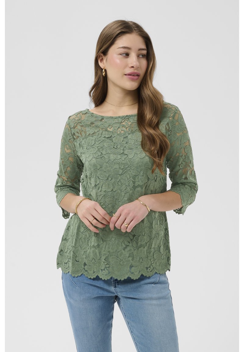 Žena s dugom valovitom kosom, noseći zelenu cvjetnu čipkastu bluzu s rukavima do tri četvrtine i plave traperice, lagano gleda u stranu.
