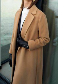 Kamelfärgad ytterrock med skräddarsydd design, sidfickor och en mjuk textur, i kombination med svarta läderhandskar och en vit topp.