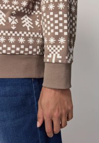 Bruine sweatshirt met een patroon en witte geometrische ontwerpen. Geribbelde manchetten en zoomdetail. Ggedragen met donkerblauwe spijkerbroeken.