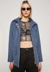Blue hooded jacket in lightweight material with front pockets, silver hardware accents, paired with a sheer patterned top and black pants.