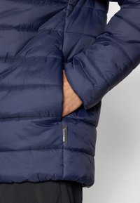 Marineblå pufferjakke med quiltet design, der har en sidelomme og et lille mærke på kanten. Blød, glat tekstur.