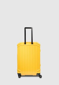Valigia rigida gialla con scanalature verticali, manico telescopico nero e quattro ruote per la mobilità. Forma semplice e rettangolare.