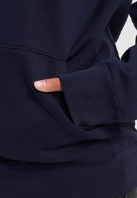 Marineblå hettegenser med ribbestruktur, frontlomme og tettsittende ermer. Stoffet ser mykt og litt elastisk ut.