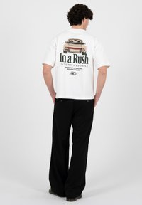 Mann trägt ein weißes T-Shirt mit einem Auto-Grafik und dem Schriftzug "In a Rush International", kombiniert mit locker sitzenden schwarzen Hosen, steht vor einem weißen Hintergrund.