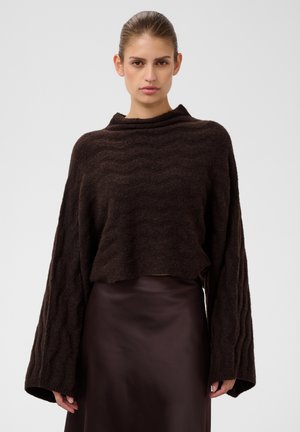 Femme portant un pull en maille lâche brun foncé à larges manches et une jupe en satin brun foncé assortie, debout devant un fond uni.