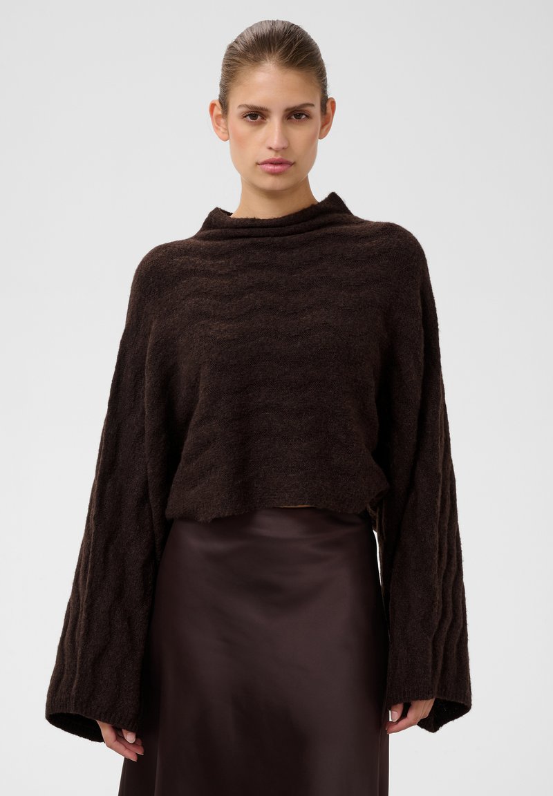 Femme portant un pull en maille lâche brun foncé à larges manches et une jupe en satin brun foncé assortie, debout devant un fond uni.