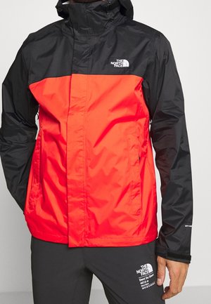 Mężczyzna noszący czarną i czerwoną wodoodporną kurtkę North Face z kapturem oraz czarne spodnie North Face z logo i napisem na udzie.