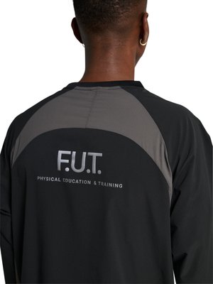 Persoon draagt een zwart-grijs jasje met de tekst "F.U.T. Physical Education & Training" op de rug, van achteren gezien.