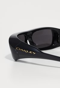 Crne sunčane naočale s sjajnom završnom obradom, s istaknutim zlatnim "Oakley" logom sa strane. Tamne leće i elegantan sportski dizajn.