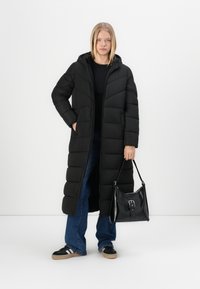 Lange zwarte pufferjas met een capuchon, met gequiltte secties. Gepaard met donkere jeans en een zwarte handtas met een zilveren hardware detail.