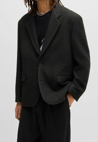 Blazer verde scuro texturizzato con chiusura a un bottone, rever a inserti e due tasche frontali. Vestibilità ampia con maniche leggermente oversize.