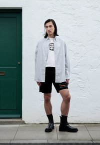 Pruhovaná košeľa na gombíky, biele tričko s potlačou, čierne šortky s logom "Rivoda" a čierne členkové čižmy. Jednoduché pozadie a zelené dvere.