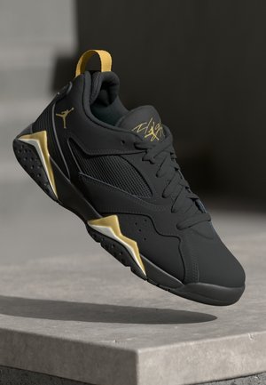 Scarpa da ginnastica nera e oro con lacci, pannello in mesh e logo distintivo, sospesa sopra una superficie in pietra grigia su sfondo scuro.