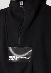Svart fleecejacka med dragkedja och hög krage. Har en glänsande svart logotypmärke "KARL LAGERFELD JEANS" och en silverfärgad dragkedja. Slät textur.