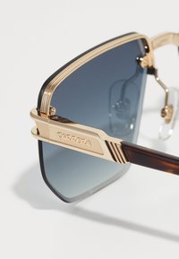 Lunettes de soleil dorées avec une monture rectangulaire, des verres teintés dégradés sombres et des accents texturés sur les branches, équipées de plaquettes de nez transparentes.