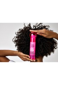 Flaska med rosa hårutjämningskräm med rundad kork och etiketten "Bed Head Tigi After Party", hålls ovanför texturerat lockigt hår.