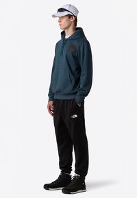 Sweat à capuche en tissu sarcelle avec poche avant et logo, associé à un pantalon de jogging noir avec un logo, et des chaussures de sport noires. Casquette blanche et lunettes de soleil.