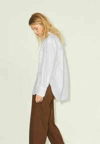 White long-sleeve shirt with a loose fit, featuring a pocket and buttoned cuffs, paired with brown wide-leg trousers. Light background.