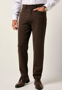 Hombre vestido con pantalones de color marrón oscuro, camisa blanca, zapatos de cuero marrón y un reloj de oro con correa de cuero marrón.