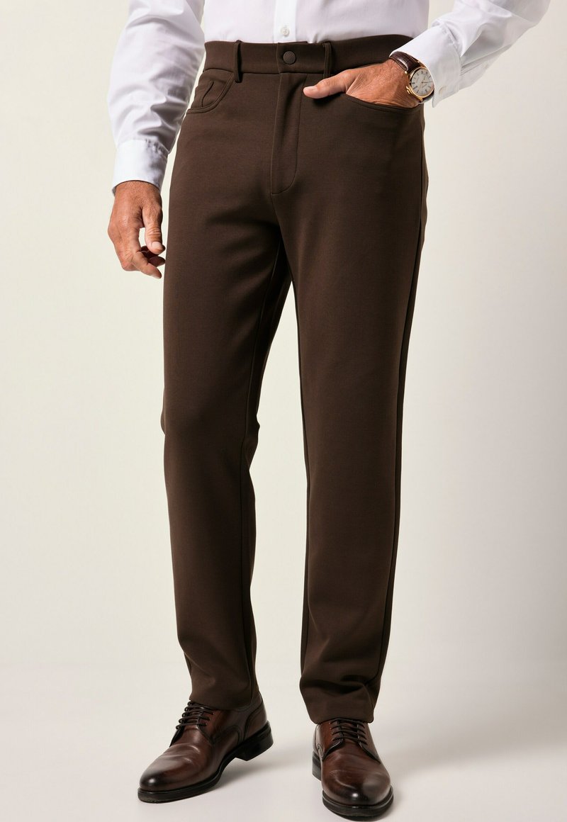 Hombre vestido con pantalones de color marrón oscuro, camisa blanca, zapatos de cuero marrón y un reloj de oro con correa de cuero marrón.