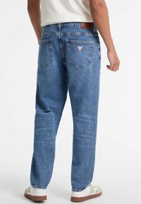 Jeans de mezclilla azul con un corte relajado, costuras visibles y dos bolsillos traseros con un parche de logo triangular. Combinados con zapatillas blancas.