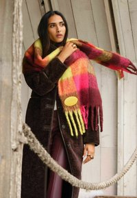 Palton pufos maro peste un top deschis, împăturit într-un fular multicolor cu nuanțe de galben, roz și roșu, având franjuri lungi. Etichetă circulară de marcă vizibilă.