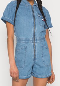 Jeansbyxdress med korta ärmar, framdragkedja, elastisk midja och två framfickor; ljusblå färg med subtil textur.