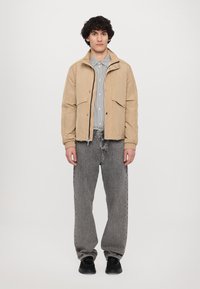 Brun jakke med frontlommer, elastiske mansjetter og glidelås, paret med en stripete skjorte og løse grå jeans. Svarte sko fullfører antrekket.