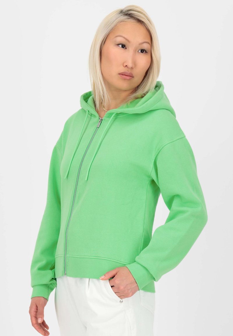 Haut de sweatshirt zippé vert en coton avec capuche, poches avant et poignets côtelés. Texture lisse et coupe moderne, associé à un pantalon blanc.