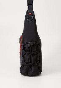 Sac bandoulière noir avec une finition texturée, présentant un dessus froncé, des sangles élastiques et une couture d'accent rouge sur un côté. Matériaux durables.