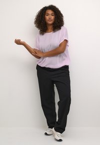 Helllila, durchsichtige Bluse mit kurzen Ärmeln, kombiniert mit schwarzen, locker sitzenden Hosen. Das Model trägt beige Sportschuhe und steht vor einem neutralen Hintergrund.