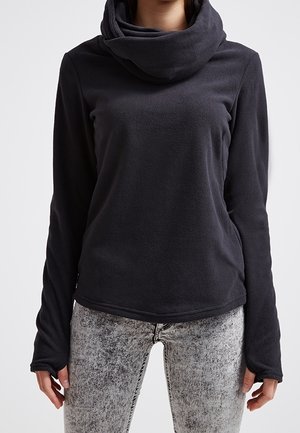 Kobieta w czarnym polarowym topie z długim rękawem, z wysokim, luźnym kołnierzem oraz szarych jeansach z efektem acid wash, stojąca na białym tle.