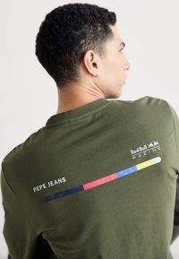 Zaļš kokvilnas T-krekls ar apaļu dekoltē, "PEPE JEANS" logo un horizontālām krāsainām svītrām sarkanā, zilā un dzeltenā krāsā uz aizmugures.