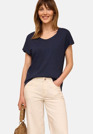 Femme souriante portant un haut bleu marine à manches courtes et un pantalon crème, tenant un sac à main beige avec une main dans sa poche.