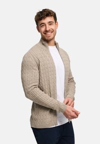 Beige strikket cardigan med glidelås og ribbestruktur, med høy krage og tettsittende mansjetter. Kombinert med en hvit T-skjorte under.