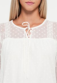Weiße Bluse mit Pünktchenstruktur, rundem Ausschnitt und Spitzenbesatz. Sie hat eine Schnürung vorne und transparente Ärmel.