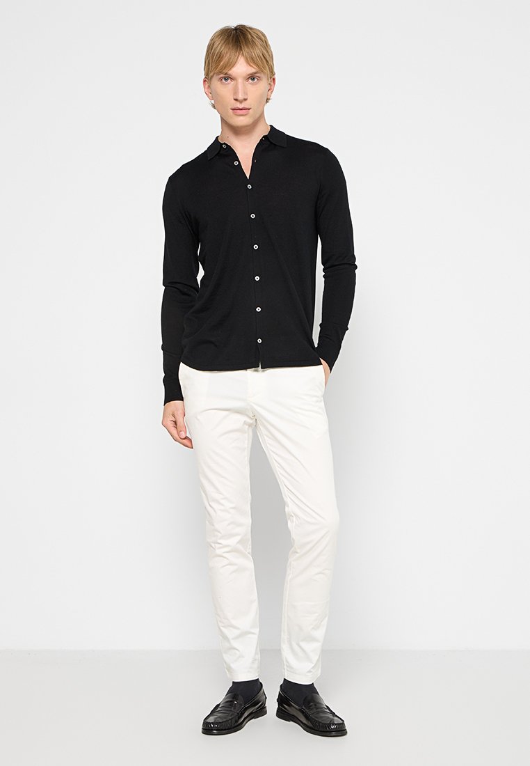 Camicia nera a maniche lunghe con colletto, texture liscia e bottoni bianchi, abbinata a pantaloni bianchi e scarpe nere.