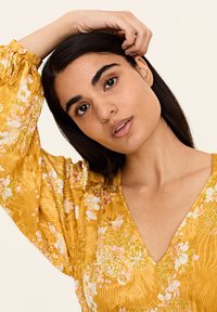 Bluză galbenă cu flori, cu mâneci lungi și ample și decolteu în V; are o țesătură texturată și modele delicate de flori roz și albe.