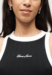 Κοντινό πλάνο ενός ατόμου που φορά ασημένια σκουλαρίκια κρίκους και μαύρο αμάνικο τοπ με λευκό περίγραμμα και κεντημένο το λογότυπο "Guess Jeans" με λευκά γράμματα.