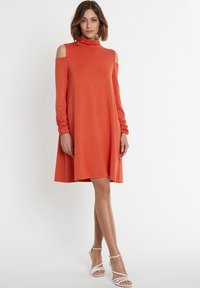 Orangekleid mit Rollkragen, kaltem Schulterdesign, langen Ärmeln und lockerer Passform; kombiniert mit weißen Riemchensandalen. Glatter Stofftextur.