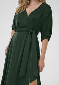 Abito a wrap verde con profondo scollo a V, maniche a sbuffo corte, vita con cinturino e spacco laterale, realizzato in un tessuto liscio e fluido.