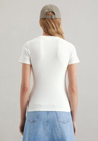 Weißes, geripptes T-Shirt mit kurzen Ärmeln, von hinten betrachtet, kombiniert mit einem hellblauen Denim-Rock. Die Trägerin hat hellbraune Haare und eine beigefarbene Kappe.