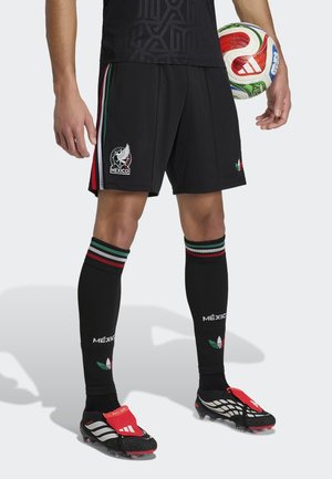 Homme portant un short de football noir du Mexique et des chaussettes avec des rayures rouges, vertes et blanches, tenant un ballon de football multicolore.
