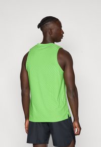 Γυναίκειο αθλητικό τοπ χωρίς μανίκια σε πράσινο χρώμα, με υφή κυμάτων, πλάγιες σχισμές και στρογγυλή λαιμόκοψη, φορεμένο με μαύρη σορτσάκι.