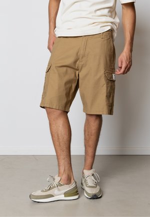 Mand iført beige cargoshorts og beige sneakers står på en lys grå gulv mod en hvid væg.