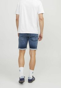 Man som bär en vit t-shirt, blå uppvikta jeansshorts, vita strumpor och marinblå sneakers, stående vänd bort mot en neutral ljus bakgrund.