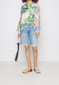 Blusa de gran tamaño, de colores y estampados; pantalones cortos de mezclilla desgastados en azul claro; mules de tejido en beige con tacón bajo; accessorios con un fleco negro.