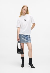Mulher com uma t-shirt gráfica branca, saia mini de denim metálico, mules pretos de salto com meias, segurando uma pequena bolsa preta contra um fundo branco.