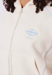 Reißverschluss-Hoodie aus leicht beigem Stoff mit blauen Akzenten. Mit einem Logo auf der Brust und geripptem Muster entlang der Nähte.